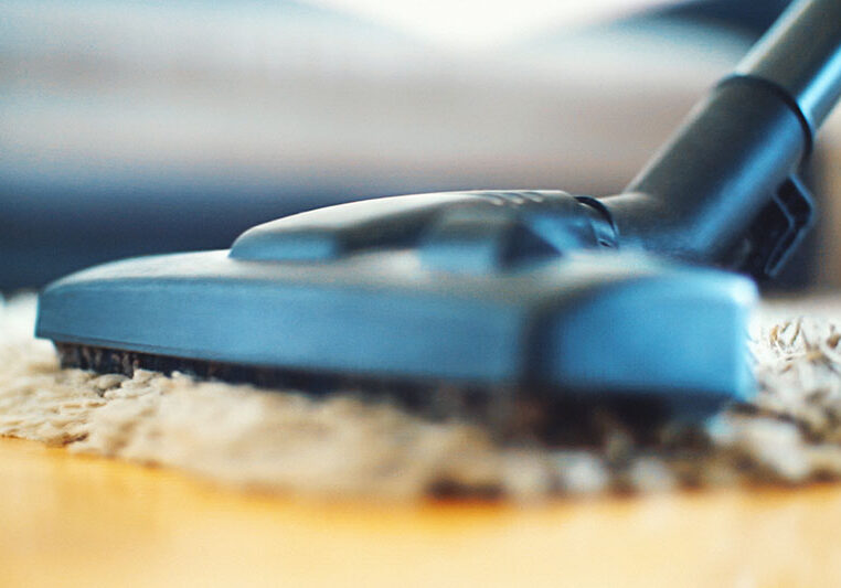Closeup low angle view of a vacuum cleaning dusting a beige living room carpet. Blurry furniture in background.